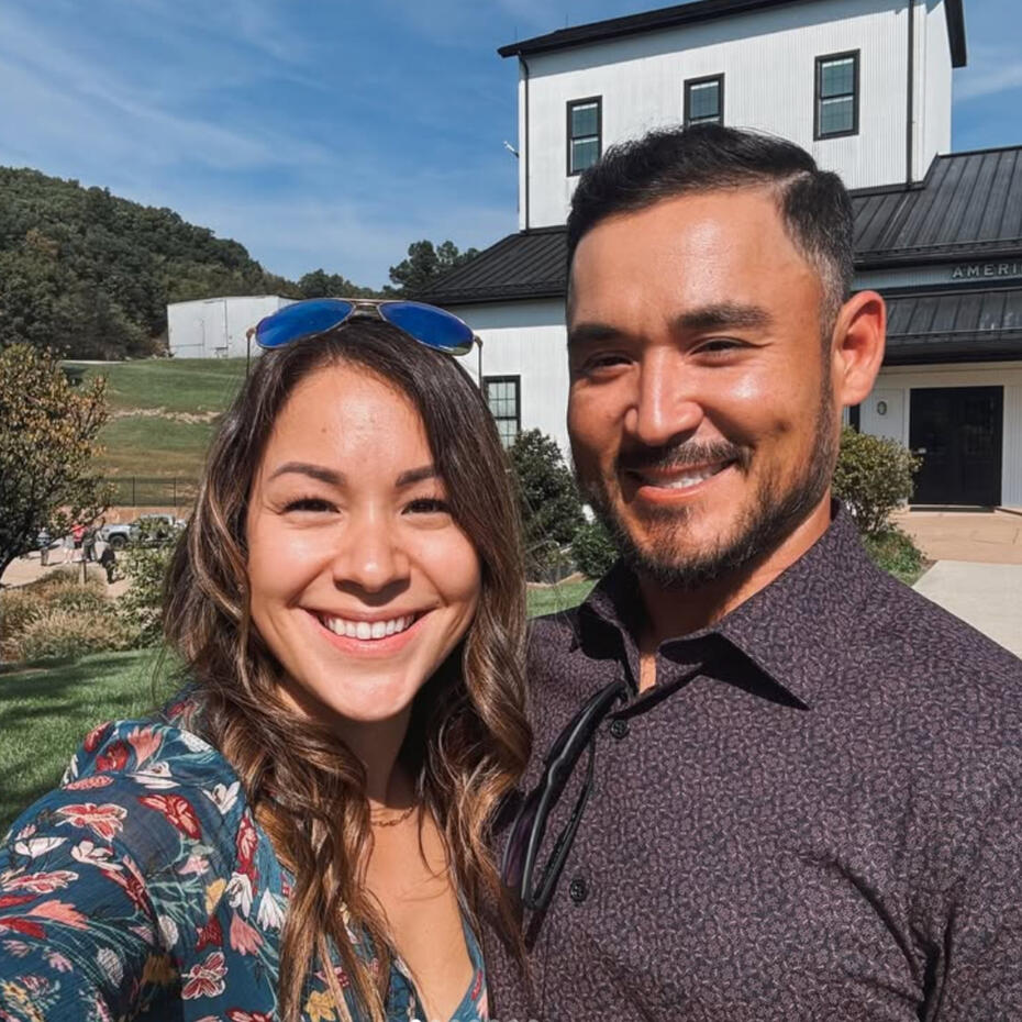 The Duvall Family A 30-something woman and man embracing and smiling in front of a two-story black and white building on a sunny day.