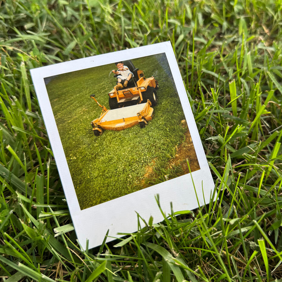Childhood Memories A polaroid photo of a young child sitting on a riding mower pretending to mow grass. The polaroid is resting on actual blades of grass.