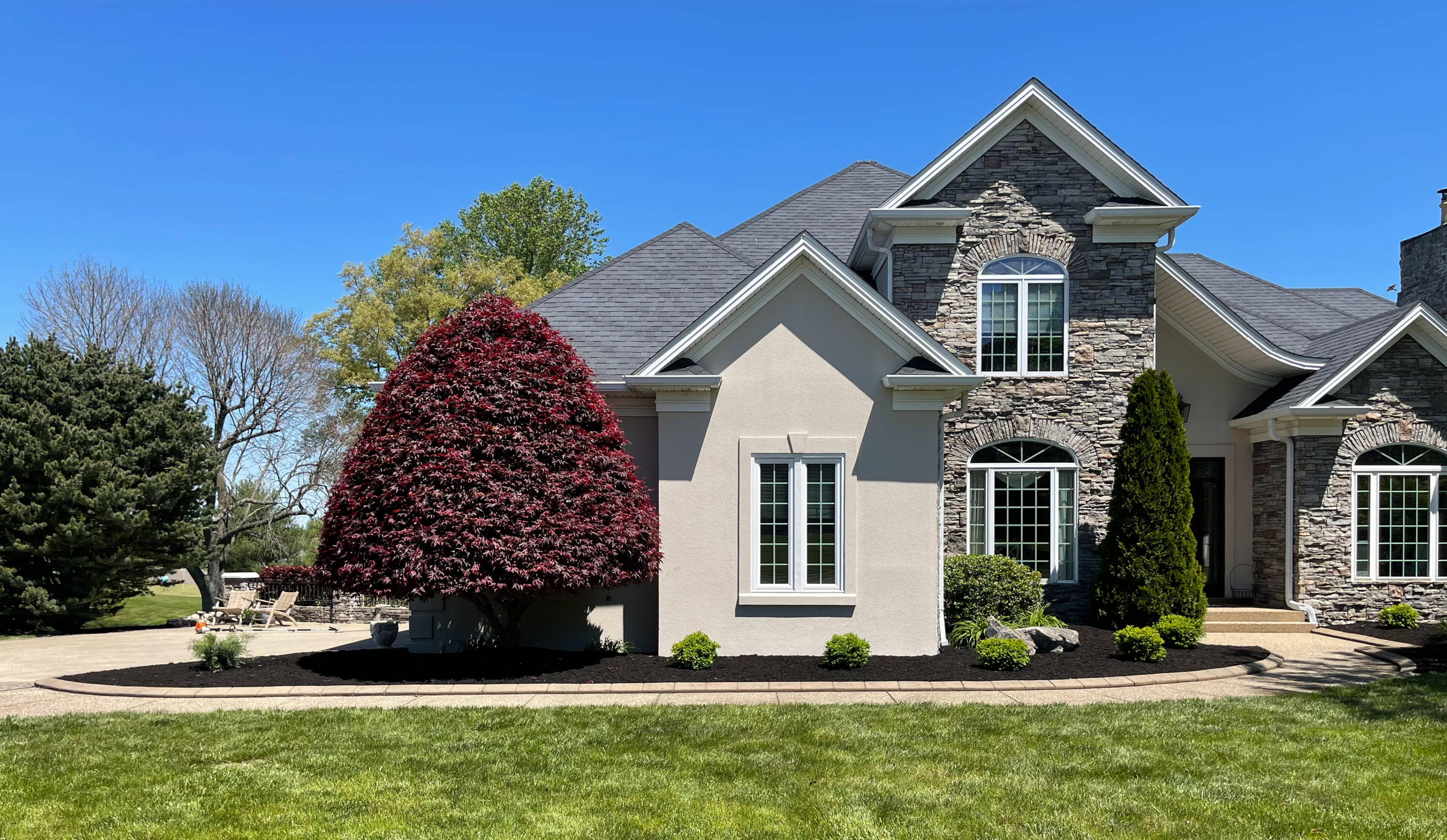 Commercial Landscaping A large, red-leafed manicured tree with triangular shape to the center left of a two-story, slate and stone home on a bright, sunny day. Some grass is visible.